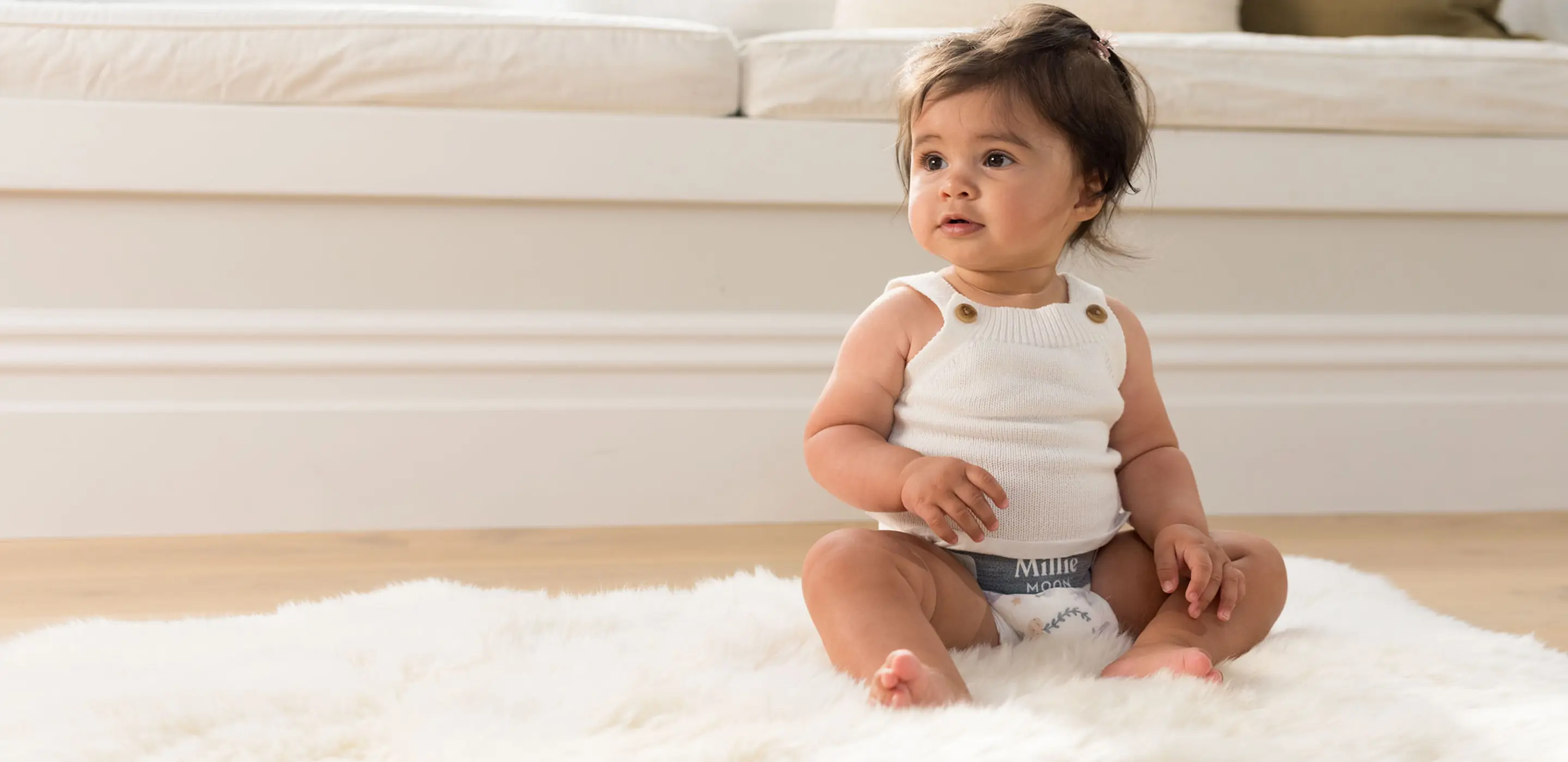 Baby in a knitted vest and a Millie Moon Luxury Diaper sitting on a sheepskin rug