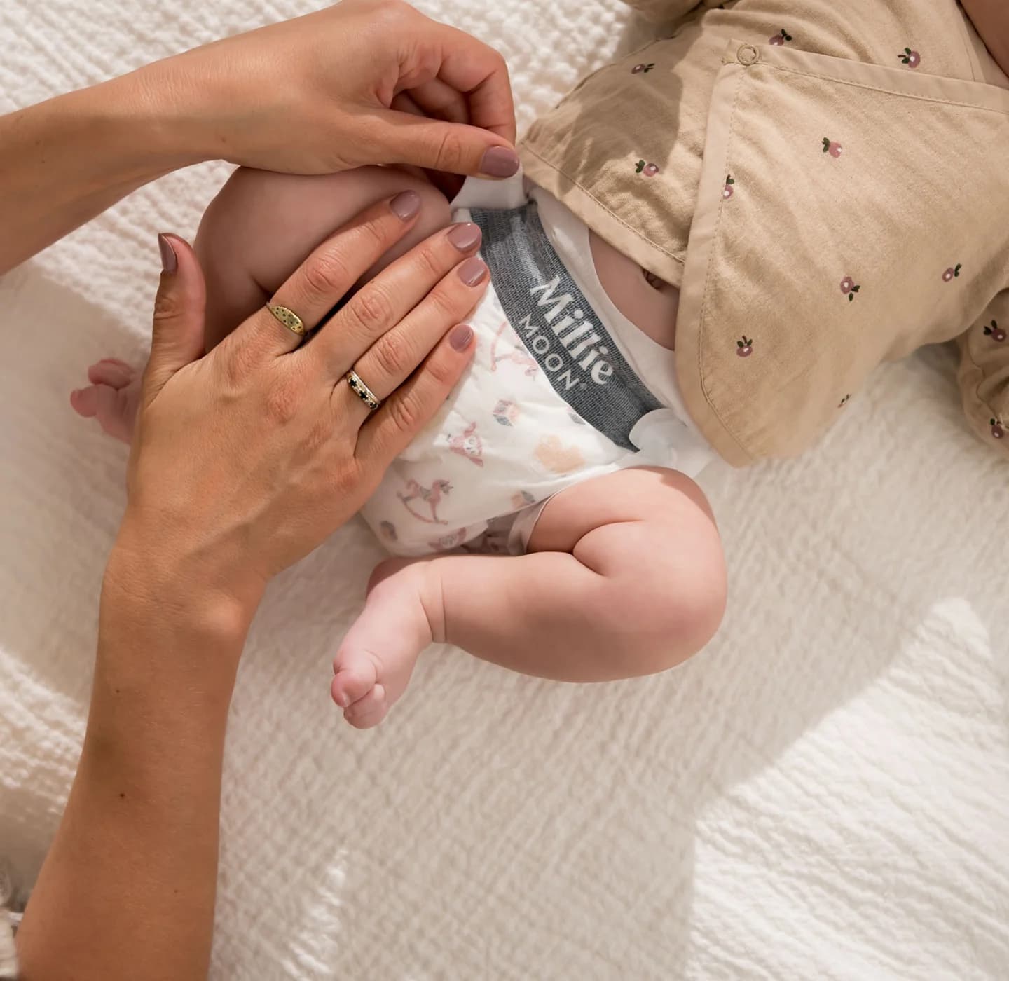 Parent fastening the waistband tab of a Millie Moon Luxury Diaper on a baby lying on a bed