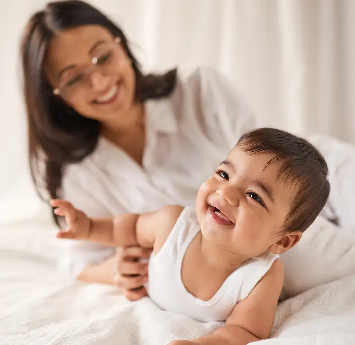 Smiling baby in a white vest lying on a bed while a parent plays alongside them